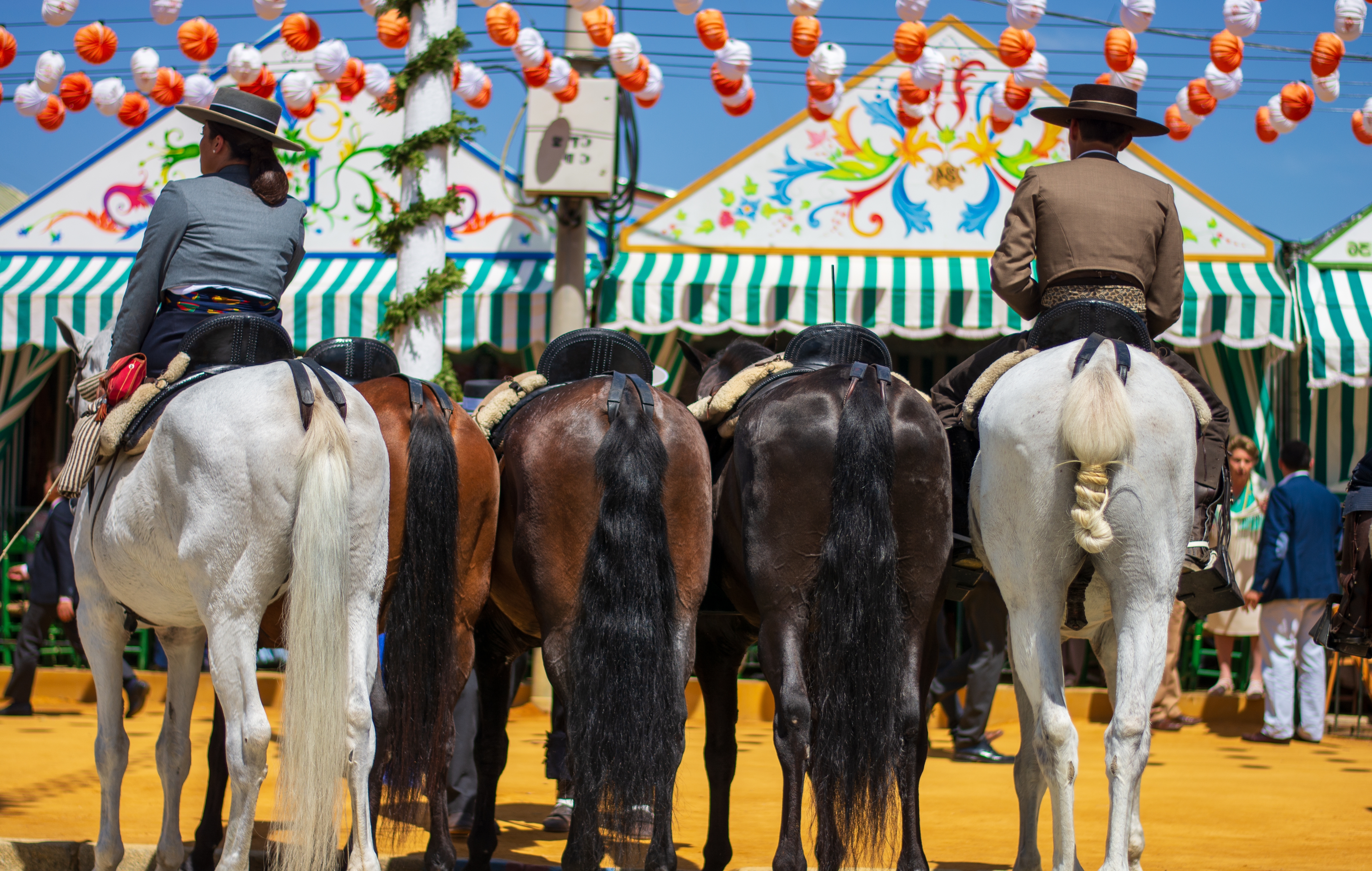 Four Andalusian horses with riders at a colorful fairground with striped tents and lanterns.